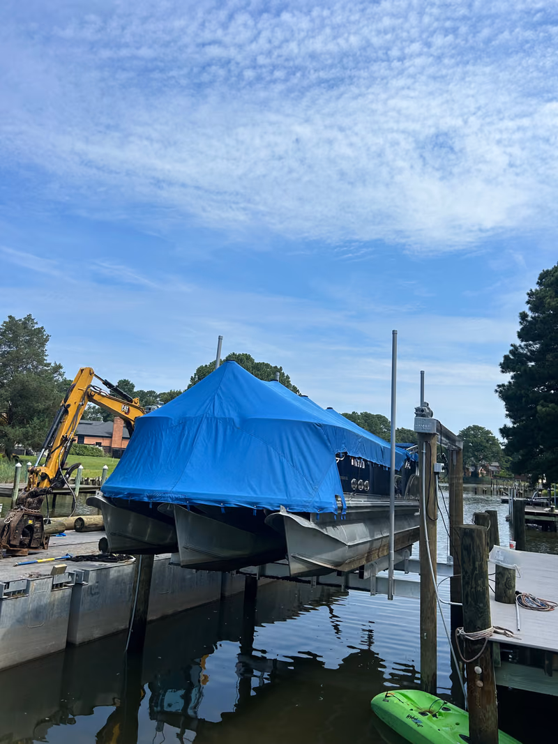 A boat under a blue cover sitting high on its boat lift, excavator on the bank behind.