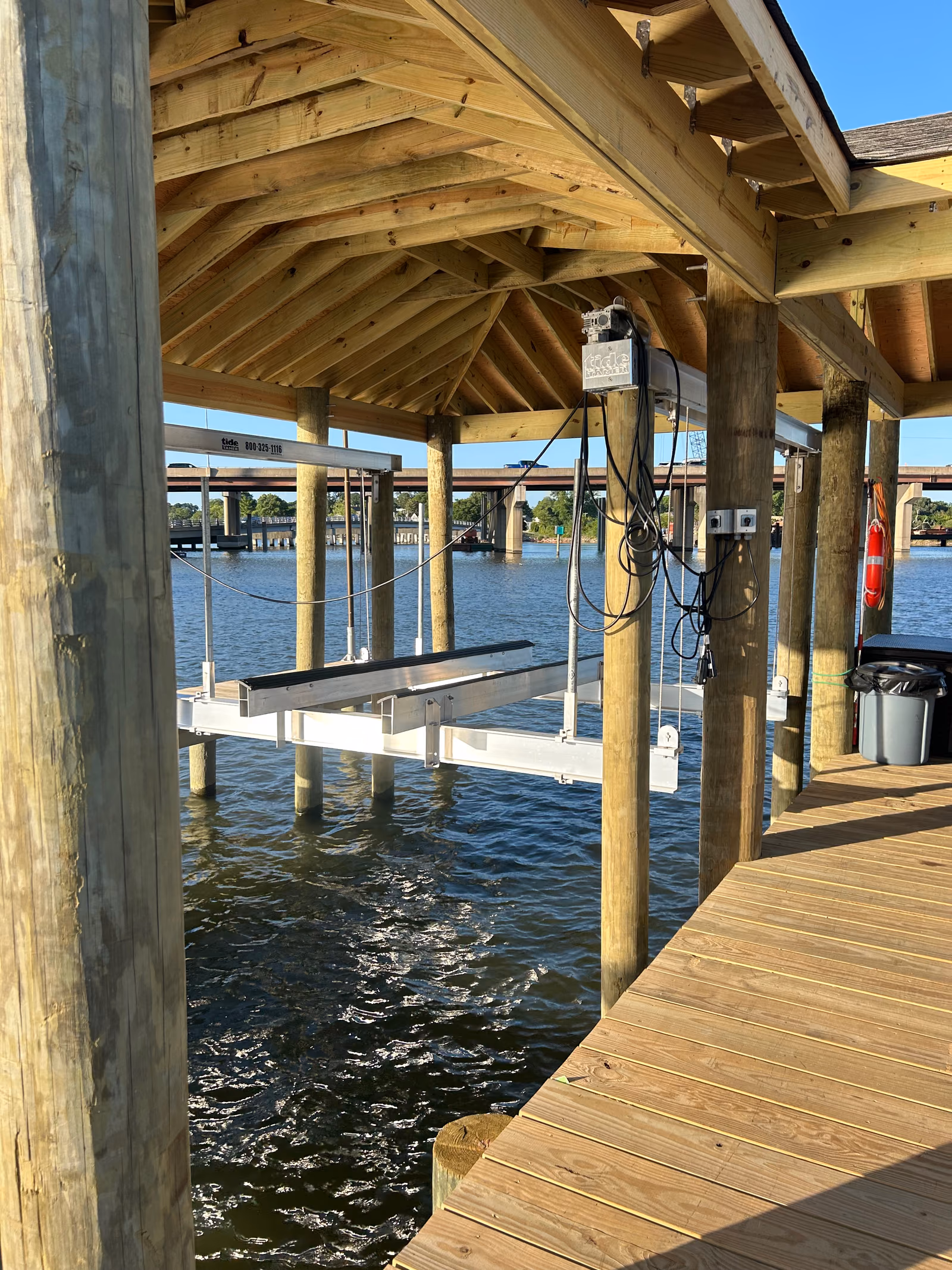 A covered boat lift inside a boathouse, looking past the cradle to a bridge beyond.