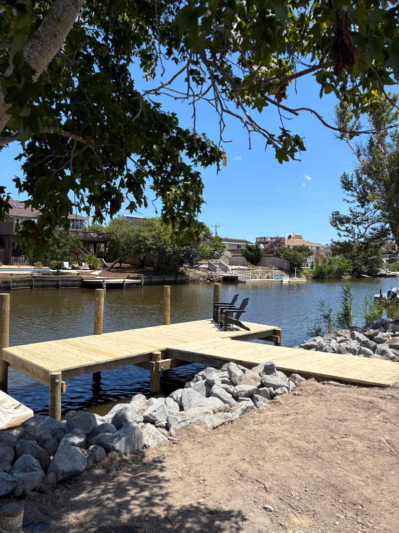 An L-shaped timber dock over a rip rap bank, two chairs on the deck in the shade.