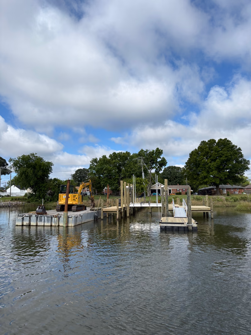 A work barge and excavator set up off a shoreline with new pilings and a float.