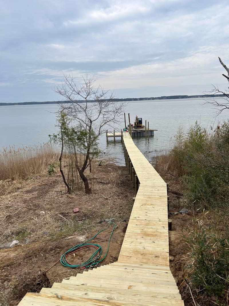 A timber walkway running down a scrub bank to a pier, work barge moored at the end.